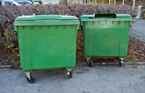 Two large green plastic wheelie bins with black wheels and lids are positioned side by side on a paved surface, likely outside a commercial or residential area in West Ham. The bins are rectangular with smooth, slightly textured surfaces and are raised slightly off the ground by four rotating wheels. The bin on the left is slightly larger and more prominent in the foreground, while the smaller bin on the right is positioned slightly behind and to the right. Behind the bins, there is a hedge with dried, brownish foliage, and a chain-link fence can be seen further back, indicating an outdoor environment. The overall scene is evenly lit, suggesting daylight conditions. These types of bins are commonly used for rubbish collection in the UK, and the setting aligns with a typical rubbish removal service context, such as those provided by Office Clearance West Ham in the local area.