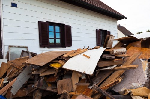 A large pile of mixed waste materials, including wooden planks, panels, and debris, is accumulated outside a white residential building with dark window shutters. The wooden pieces vary in size, color, and material, with some showing weathered textures and others smoother finishes. The debris is situated on a paved surface close to the house, slightly spilling onto the driveway or garden area. In the background, the building's exterior wall and a portion of the roof are visible, indicating a suburban setting likely in West Ham. The scene appears to be part of a rubbish removal or clearance process, with no visible activity or tools in use. The overall area is illuminated by natural daylight, contributing to a clear and neutral visual impression suitable for documenting waste removal services conducted by Office Clearance West Ham.