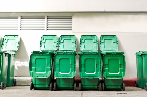 A row of eight green wheeled rubbish bins with closed lids, positioned on a paved outdoor surface in front of a white building wall featuring vent panels near the top. The bins are aligned in two evenly spaced groups, with the front row slightly closer to the camera and the back row slightly behind, all placed along the edge of the pavement. The environment appears to be an exterior waste collection point, possibly in West Ham, with the bins suitable for refuse or recyclables. The bins have textured plastic surfaces with smooth, raised rectangular lids and small wheels at the base for mobility, all uniformly in a bright green colour. The wall background is clean with a red-painted curb edge visible at the bottom, and there is natural daylight illuminating the scene, remaining neutral and professional in tone, illustrating typical rubbish collection infrastructure managed by Office Clearance West Ham.
