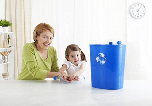 A woman with short blonde hair, dressed in a light green top, is seated at a white table alongside a young girl with brown hair styled in pigtails, wearing a white shirt. They are smiling and appear to be engaging with a clear plastic bag of waste or recyclable materials placed in front of them. To the right, there is a blue recycling bin with a white recycling symbol on the front, positioned on the same white surface. The background shows a modern, bright interior environment with white walls, a portion of a clock on the upper right, and some home decor elements. The scene likely depicts a household activity related to rubbish sorting or recycling as part of waste management services offered by Office Clearance West Ham, with an emphasis on environmentally responsible disposal practices within the area around East London.