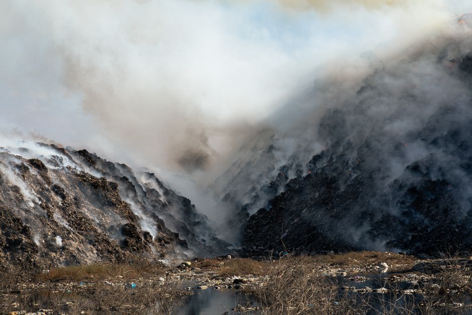 An expansive landfill site filled with a large volume of mixed waste materials, including plastic bags, packaging, and household debris, spread across a sloped terrain of compacted gravel and dirt. In