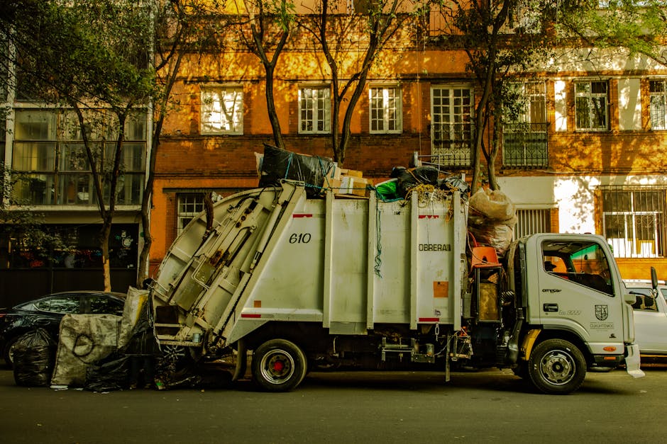 The image displays a collection of various discarded items on a paved driveway or street surface, in front of a residential building with brick walls and a wooden fence. In the foreground, there are l
