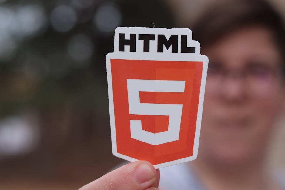A person holds a cut-out sticker featuring the HTML5 logo, which consists of a shield shape with a white border and an orange background, displaying a large white number '5' in the center and the lett